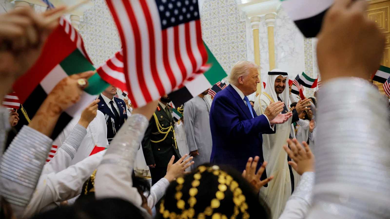 A crowd waves U.S. and UAE flags around a central figure in a blue suit, surrounded by officials and locals in traditional attire, conveying a festive atmosphere.